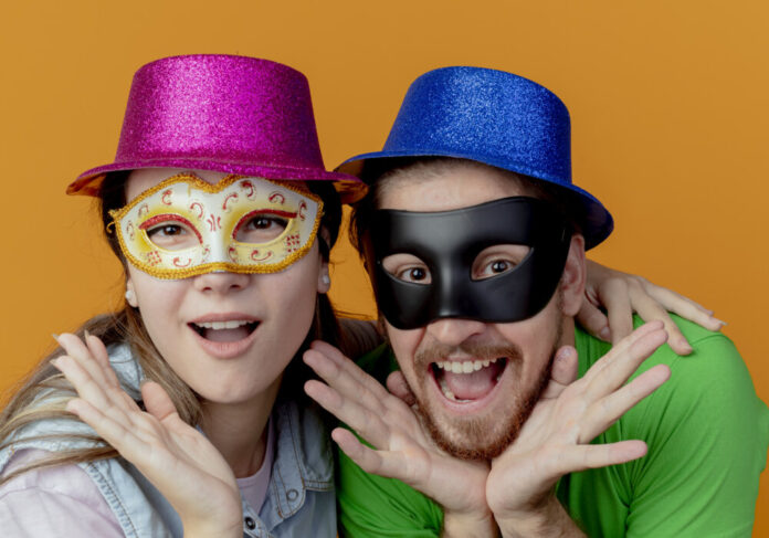 young excited couple wearing pink and blue hats put on masquerade eye masks putting hands on chin isolated on orange background with copy space